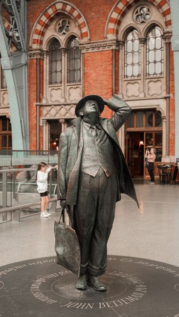 A bronze statue of John Betjeman, depicted as a middle-aged man in a suit, waistcoat, and overcoat, is positioned inside a historic train station with high arched windows and red brick walls. The stat