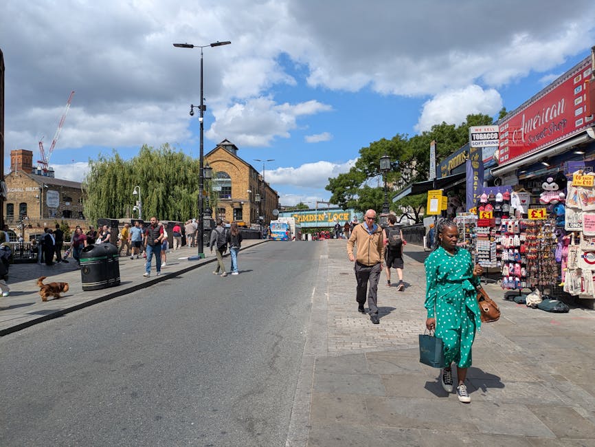 A busy outdoor street scene at Camden Market during daytime featuring a paved sidewalk and a slightly inclined asphalt road. The sidewalk is clean and well-maintained with light-colored paving stones, alongside a row of small shops and market stalls on the right, displaying colorful merchandise including clothing, accessories, and souvenirs. The shops have vibrant signage, and some items are hung on display racks and hooks. Pedestrians, including a woman in a turquoise polka-dot outfit carrying a shopping bag and a brown handbag, are walking on the sidewalk, while others stand browsing the stalls. A man in a light brown jacket, black trousers, and sunglasses walks past, and a dog lies on the street, close to a black trash can. In the background, there are taller brick buildings with large glass windows, under a partly cloudy sky with scattered white clouds and bright sunlight illuminating the scene. The overall environment appears tidy, lively, and well-maintained, reflecting a typical bustling market street, with Camden Cleaner noted as the service provider for street and commercial cleaning.