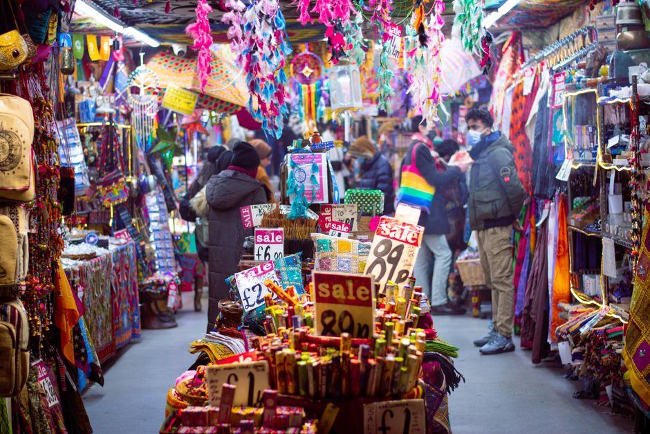 An indoor market stall at Camden Market bustling with shoppers and vibrant displays of colorful fabrics, accessories, and decorative items. The stall features various textiles, jewelry, and merchandise organized on shelves and tables, with signs indicating sale prices such as £6, £2, and 89p. Ceiling decorations include hanging paper and fabric ornaments in bright hues, creating a lively atmosphere. Customers are browsing, some wearing winter coats and masks, with a mix of natural and artificial lighting illuminating the diverse array of products. Camden Cleaner’s deep cleaning service ensures the cleanliness and hygiene of such busy commercial spaces, maintaining surfaces like floors, counters, and display areas in pristine condition to support safe shopping environments and high standards of hygiene in Camden Market. The scene captures the vibrant, cluttered ambiance typical of market stalls focused on retail and customer engagement.