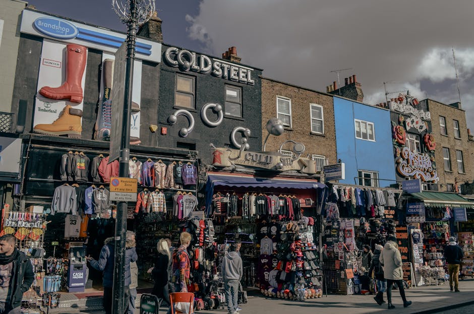 A busy outdoor street scene at Camden Market during daytime featuring a paved sidewalk and a slightly inclined asphalt road. The sidewalk is clean and well-maintained with light-colored paving stones, alongside a row of small shops and market stalls on the right, displaying colorful merchandise including clothing, accessories, and souvenirs. The shops have vibrant signage, and some items are hung on display racks and hooks. Pedestrians, including a woman in a turquoise polka-dot outfit carrying a shopping bag and a brown handbag, are walking on the sidewalk, while others stand browsing the stalls. A man in a light brown jacket, black trousers, and sunglasses walks past, and a dog lies on the street, close to a black trash can. In the background, there are taller brick buildings with large glass windows, under a partly cloudy sky with scattered white clouds and bright sunlight illuminating the scene. The overall environment appears tidy, lively, and well-maintained, reflecting a typical bustling market street, with Camden Cleaner noted as the service provider for street and commercial cleaning.