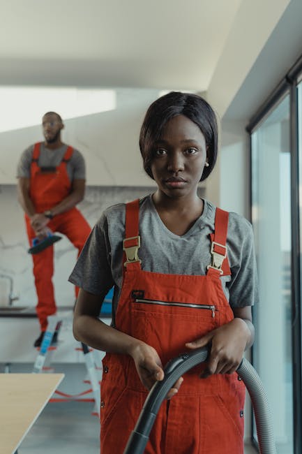 A professional cleaner, a woman with dark skin and short black hair, is standing on a modern, light-colored tiled floor inside a residential space with large glass sliding doors. She is wearing an ora